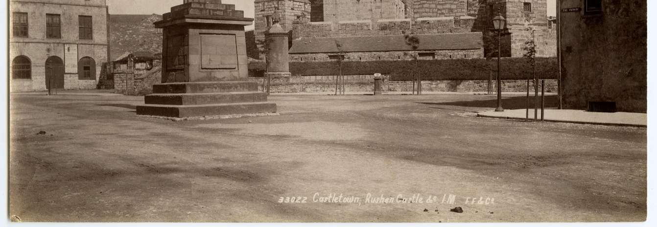 A vintage sepia-toned photograph depicting a stone war memorial or monument situated in a town square with historic stone buildings and Rushen Castle visible in the background.