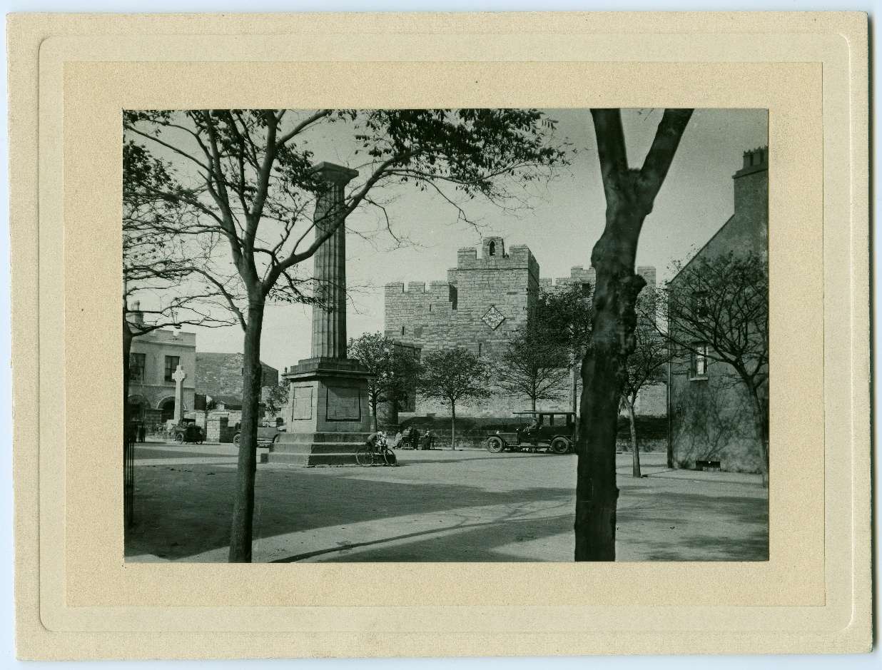 A black and white historical photograph showing a stone tower or castle structure in the background with a tall column monument in the foreground.