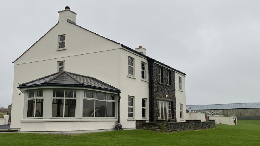 A photograph of a white two-story detached house featuring a stone-clad section and a large bay window, set in a grassy rural area with a long agricultural building in the background.