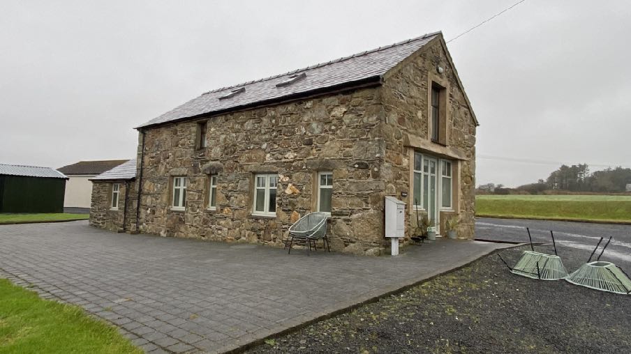 A photograph of a detached stone building with a slate roof and paved driveway, situated in a rural setting with a green outbuilding to the side.