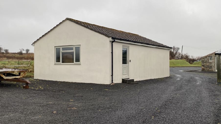 A photograph of a white, single-storey building with a tiled roof and gravel driveway in a rural setting.