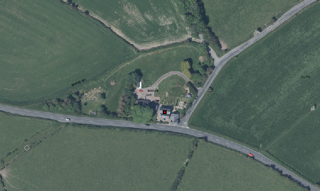 Aerial satellite image showing a detached rural property with a driveway, surrounded by green fields and hedgerows.