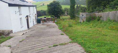 A photograph showing the exterior of a white building with a paved driveway and a grassy area, set against a rural backdrop with green hills.