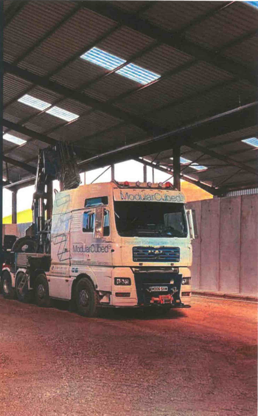 A photograph showing a large white truck parked inside a large open-sided industrial shed or warehouse structure with a metal roof.