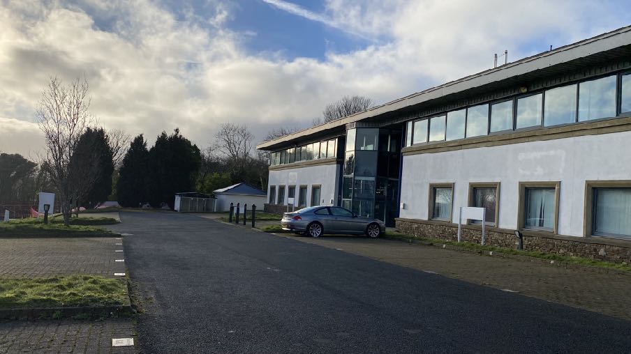 A photograph showing the exterior of a white, single-story commercial building with large windows, likely an office block, with a car parked on the paved driveway.