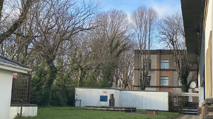 A photograph showing the exterior of a multi-story building surrounded by trees and a white boundary wall, likely the subject of the office conversion application.