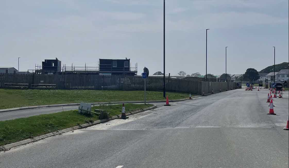 A photograph showing a paved road with traffic cones and a wooden hoarding fence, behind which are temporary site cabins or structures.