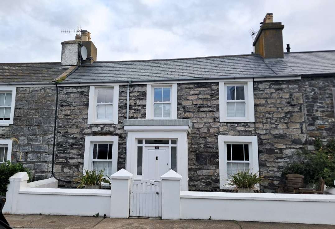 A street-level photograph of a two-story stone terrace house featuring white sash windows, a white front door, and a rendered boundary wall.