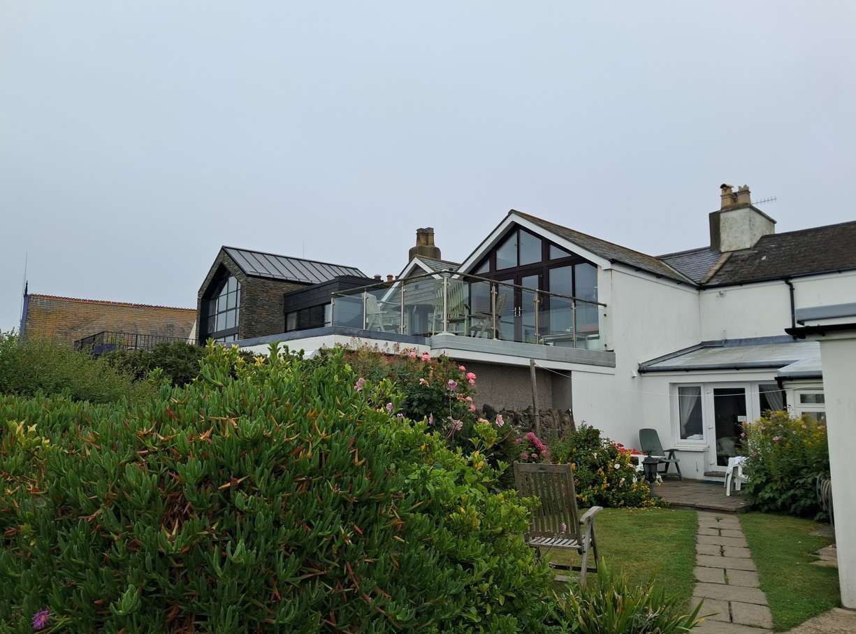 A photograph showing a residential property with a large, modern two-story extension featuring a glazed terrace and balcony area.