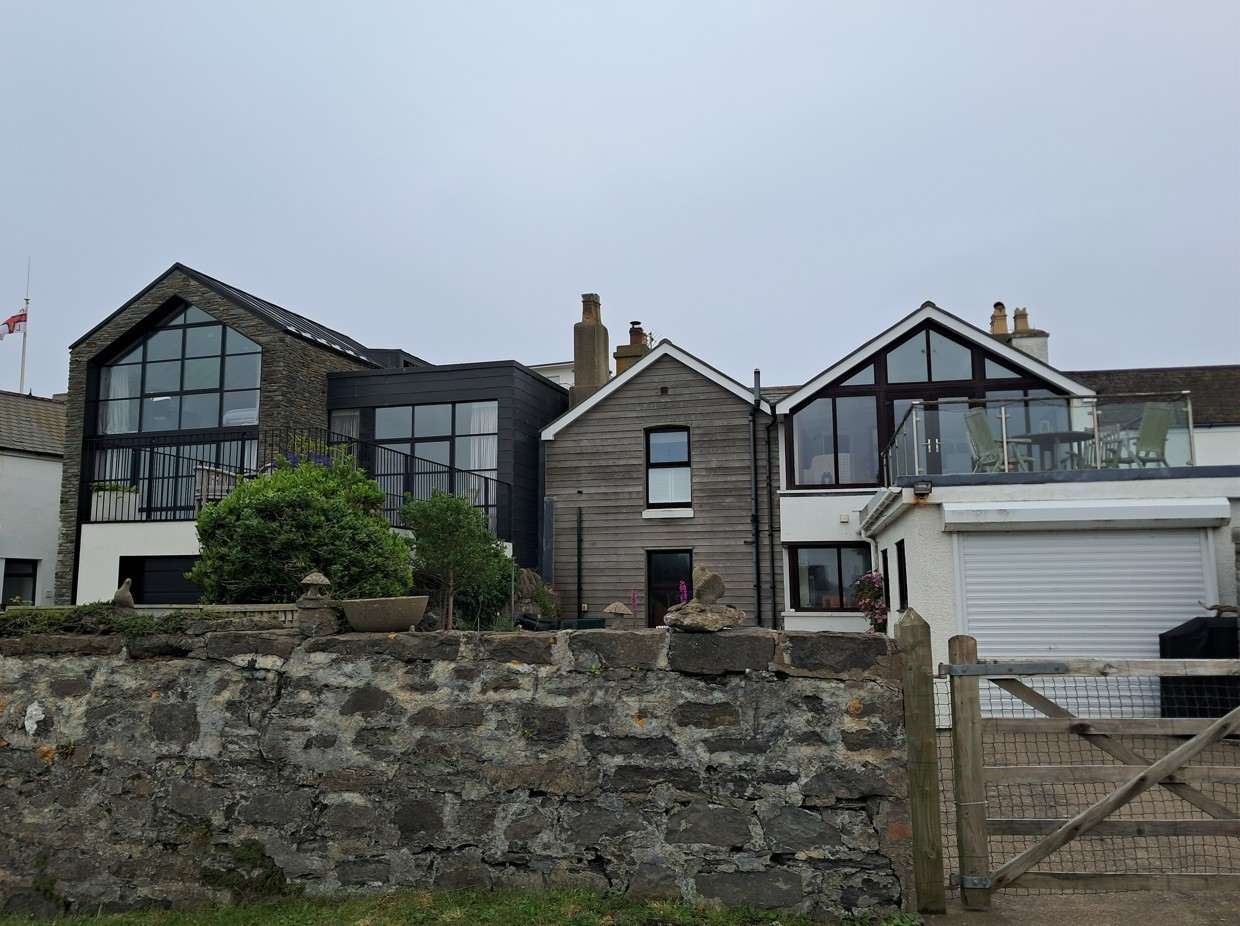 A street-level photograph showing a row of residential properties, including a modern stone-fronted house and a property with a garage and terrace, viewed from behind a stone wall.