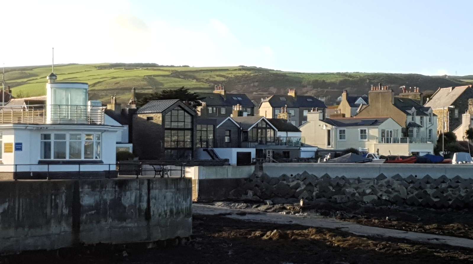 A photograph showing a row of residential houses along a coastal promenade, featuring a white house with a glass extension and a dark stone house with a large conservatory, set against a backdrop of green hills.