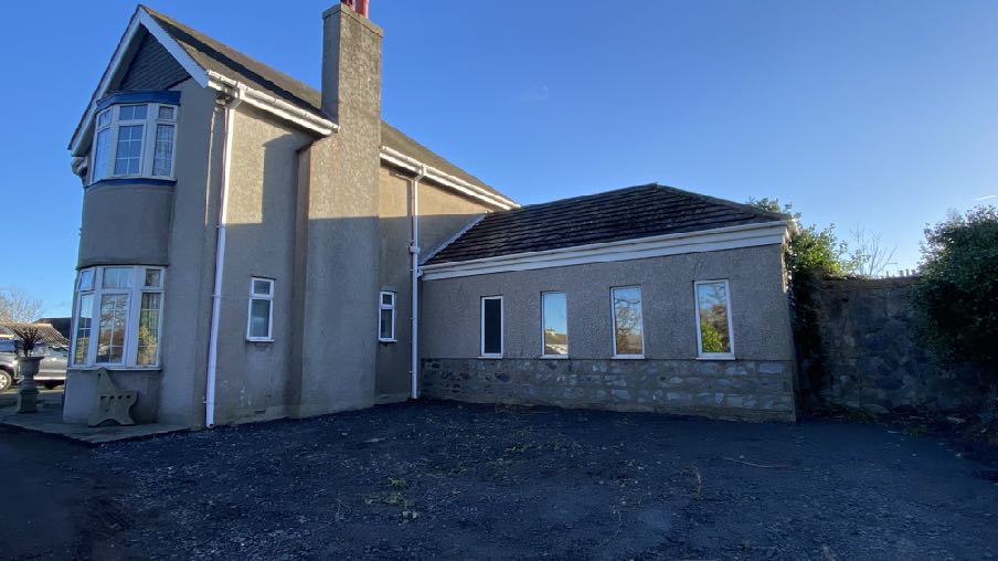 A photograph showing the rear elevation of a detached house featuring a two-story section with a bay window and an attached single-story extension with a stone plinth and gravel driveway.