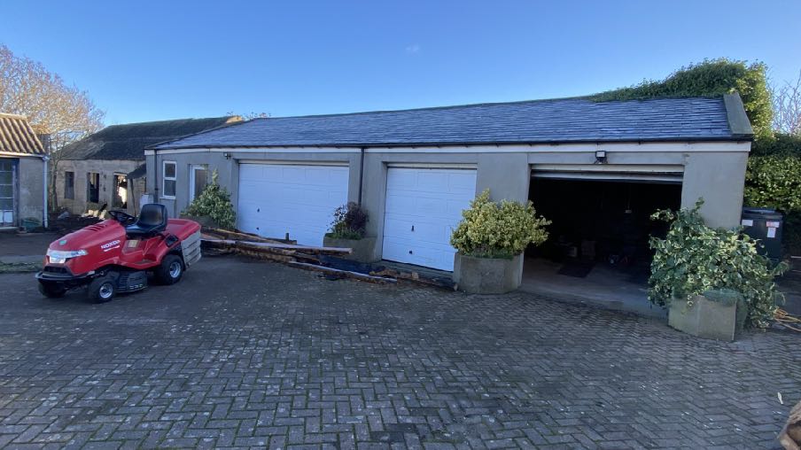 A photograph showing a paved driveway with a red lawnmower in front of a single-story building featuring white roller garage doors and potted plants.