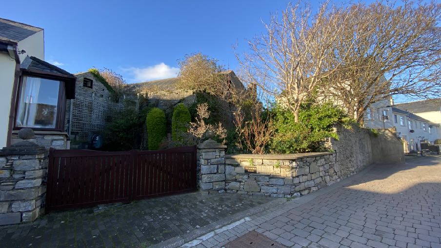 A street-level photograph showing a residential property with stone boundary walls, a wooden gate, and surrounding vegetation under a clear blue sky.