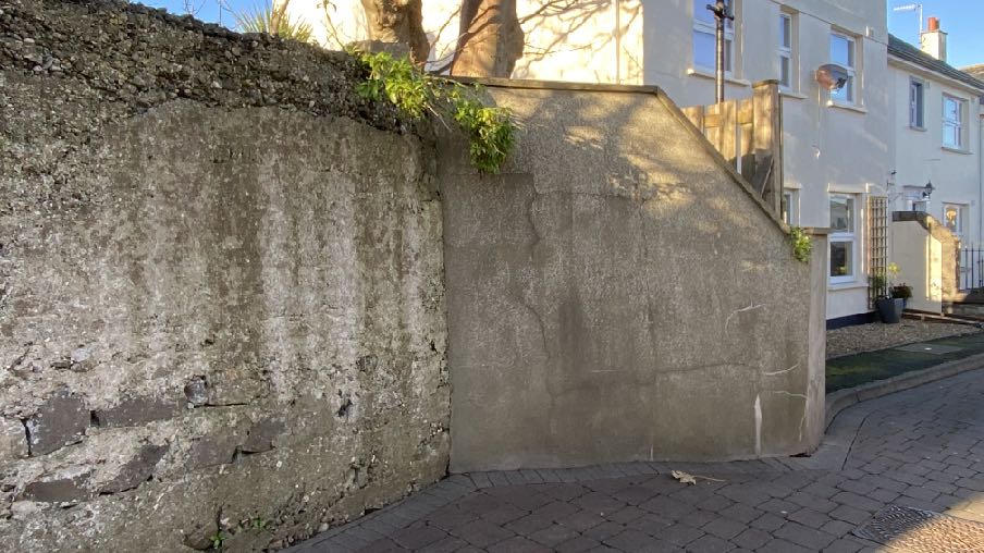 A photograph showing a boundary wall with rough and smooth rendered sections, with a white residential building visible in the background.