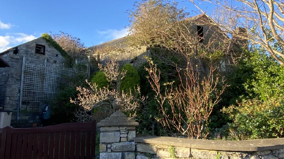 A photograph showing a stone building with a slate roof, partially obscured by trees, viewed from behind a stone wall and wooden gate.
