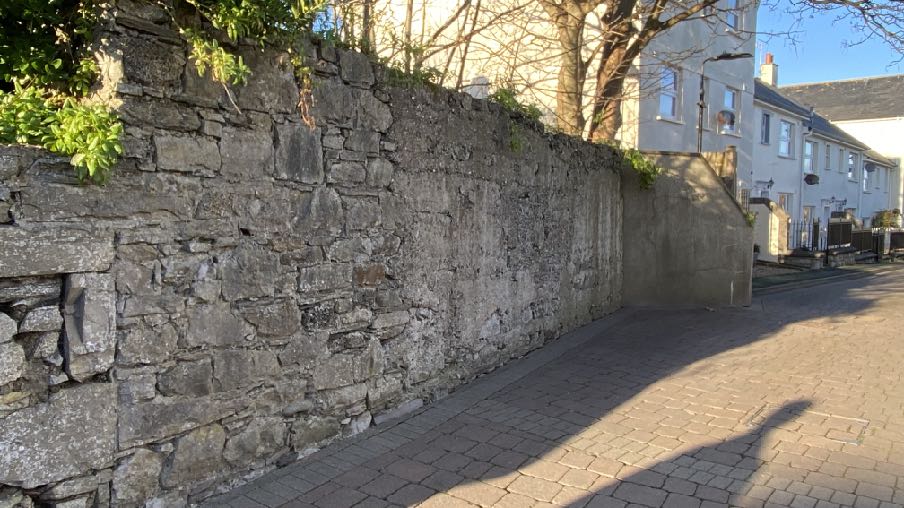A photograph of a long, weathered stone boundary wall running alongside a paved street, with residential buildings visible in the background.