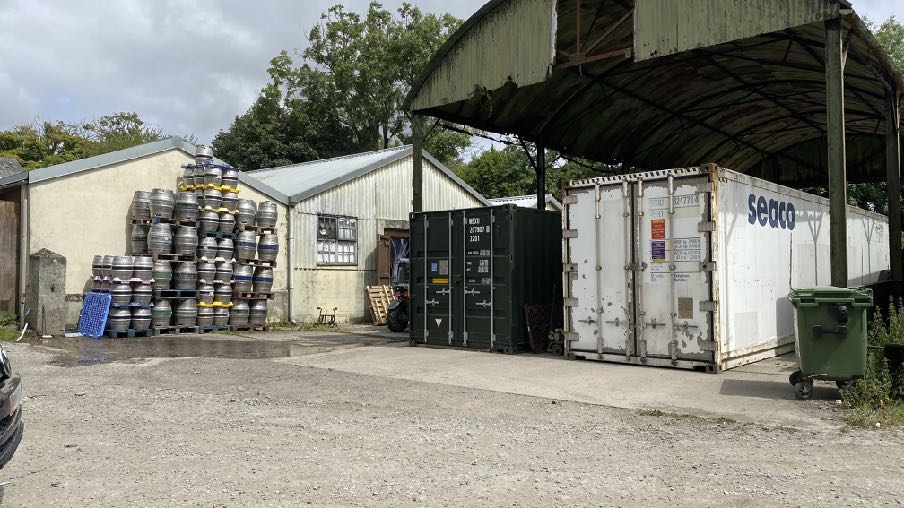 A photograph of an outdoor commercial storage yard featuring stacked beer kegs, a white warehouse building, and shipping containers under a metal shelter.