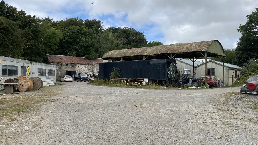 A photograph of a rural industrial or agricultural site featuring a gravel yard, a stone building, and a large open-sided metal shed structure.