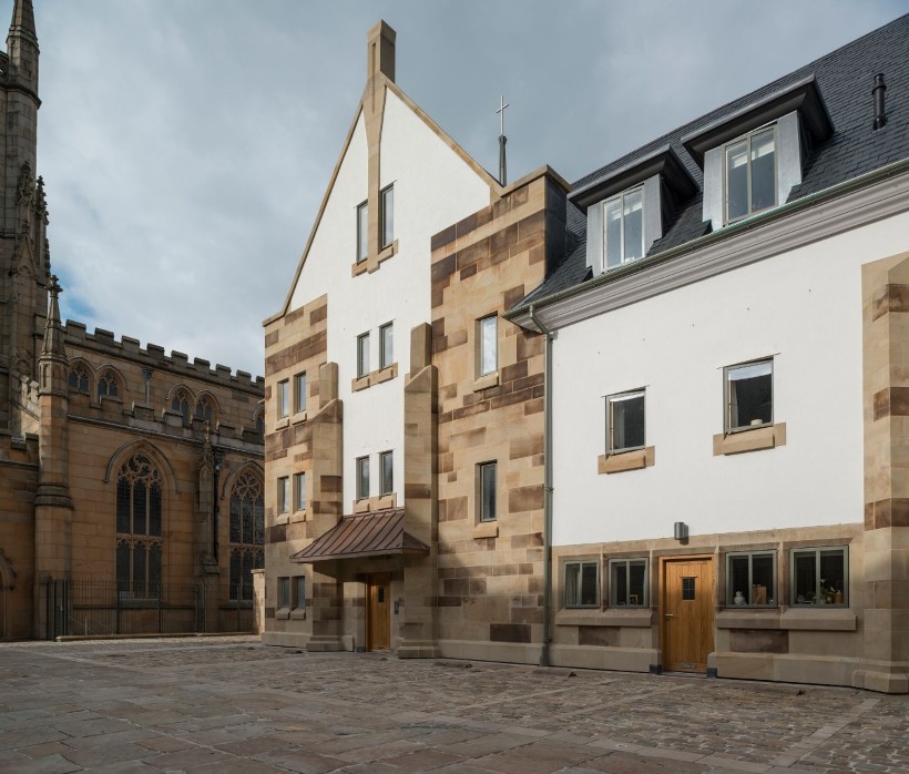 This image displays an exterior view of a white building with stone quoins and dormer windows, situated in a paved courtyard adjacent to a large Gothic-style stone church.