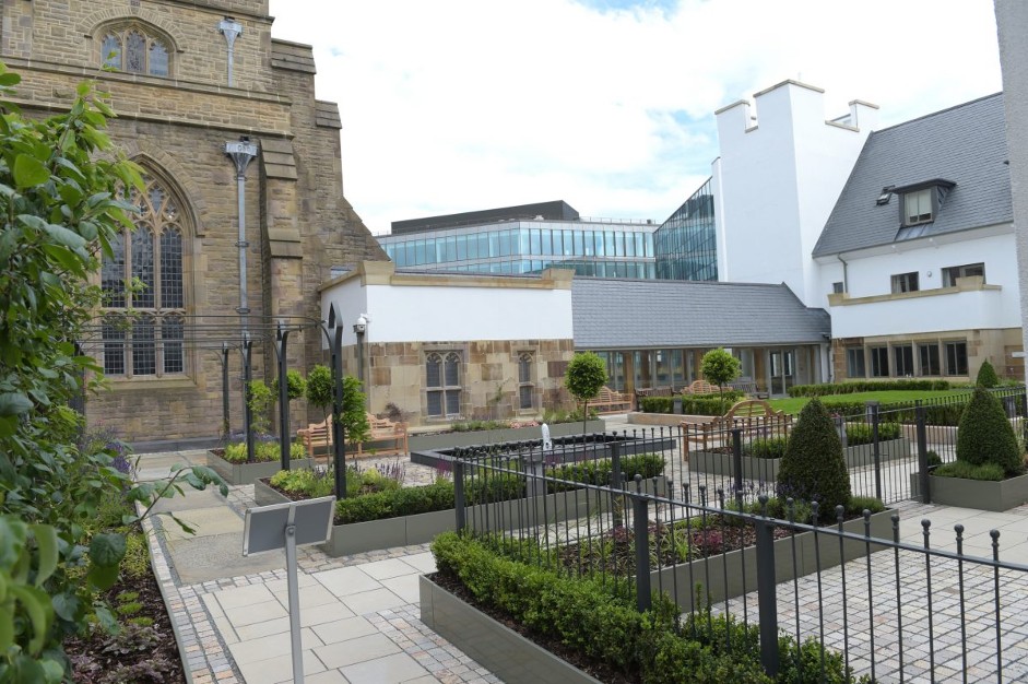 A photograph showing a landscaped courtyard with paved paths and planters in the foreground, adjacent to a historic stone building with a large arched window on the left and a modern white residential building on the...