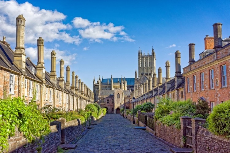 A photograph of a historic cobblestone street lined with stone buildings and numerous chimneys, leading towards a large cathedral in the distance.