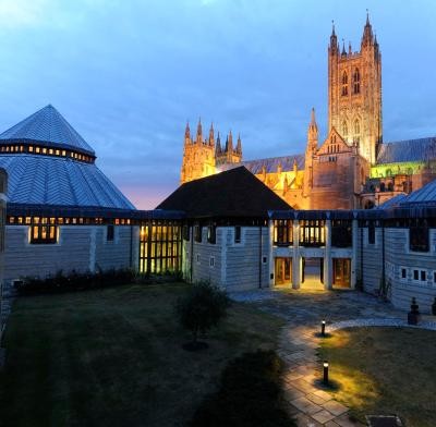A twilight photograph of a courtyard featuring stone buildings with modern extensions and a large illuminated cathedral tower in the background.