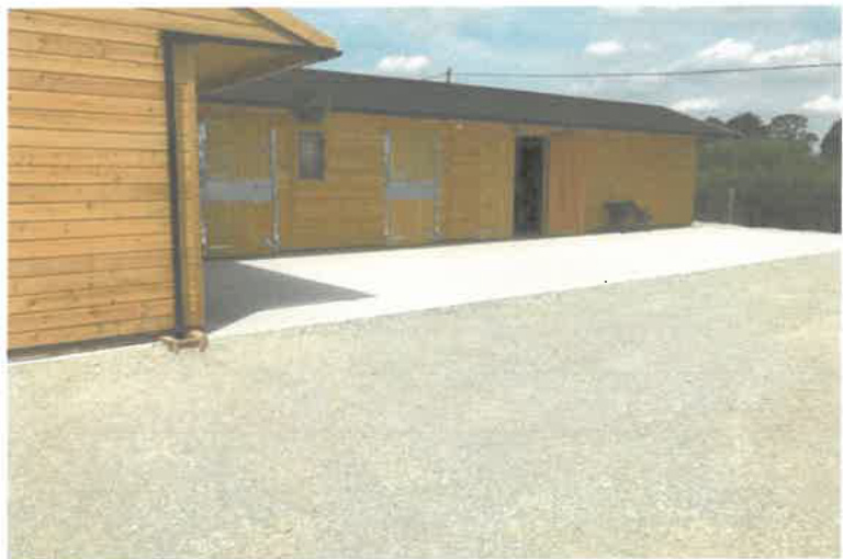 A photograph showing a wooden agricultural building or stable with large doors, situated next to a gravel yard.