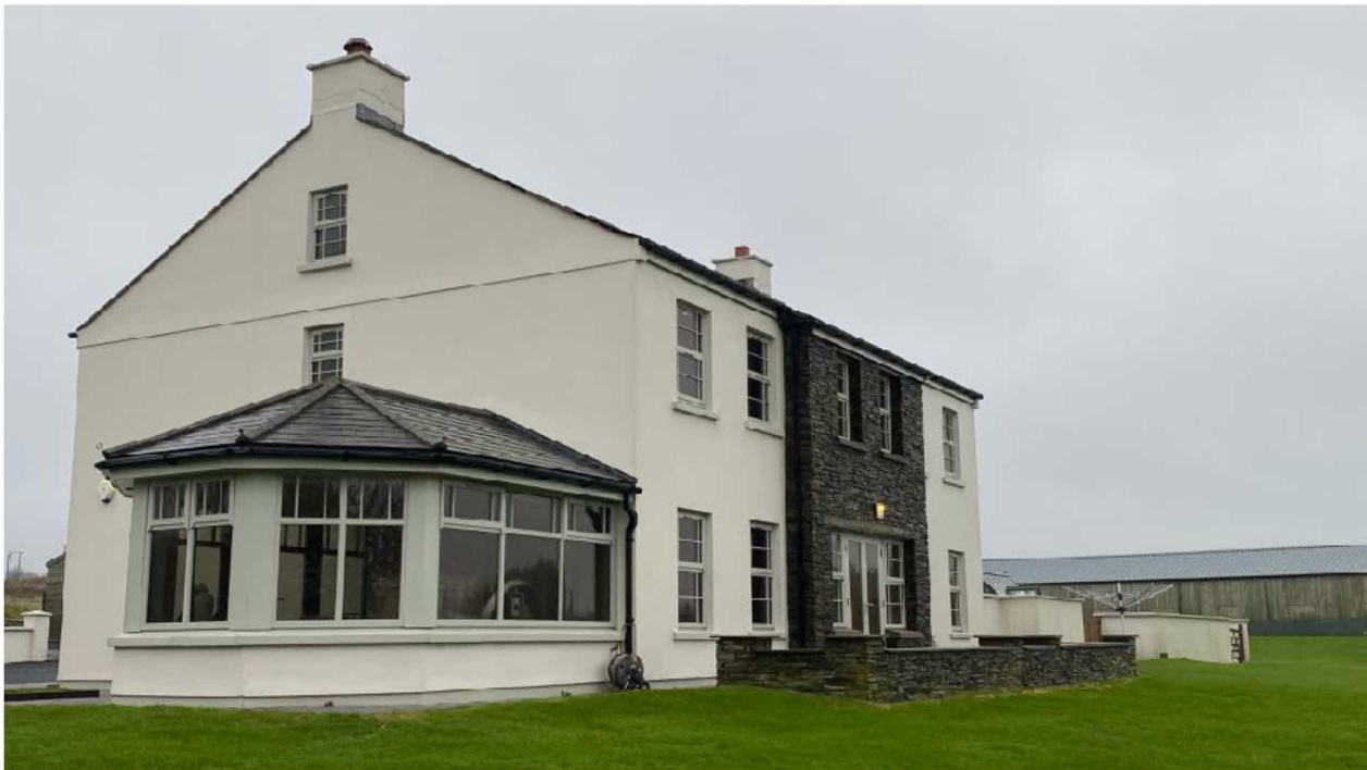 A photograph of a large white detached house featuring a prominent bay window extension and a stone facade section, set in a rural setting with an agricultural building in the background.