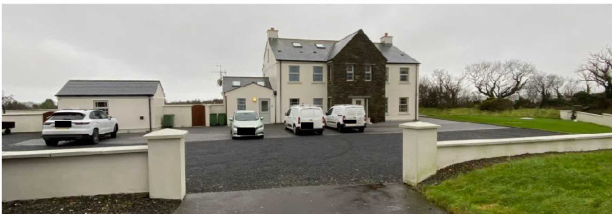A photograph showing a large detached house with a stone extension and a gravel driveway containing several vehicles. A smaller outbuilding is visible to the left and a grassy field is on the right.
