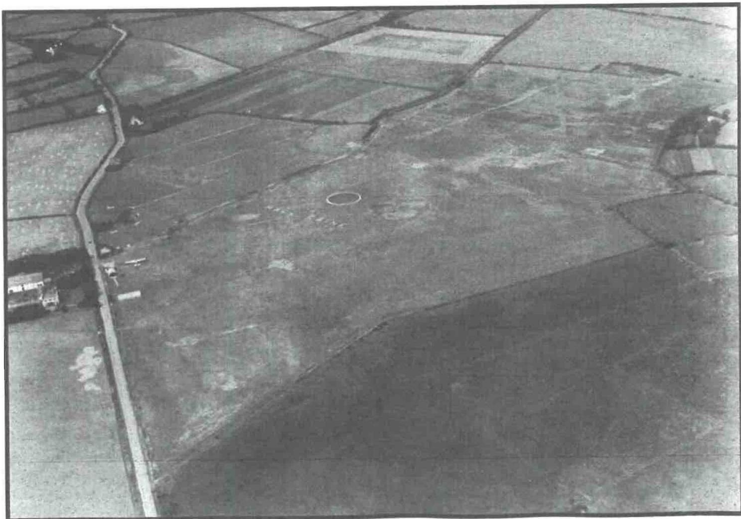 A grainy black and white aerial photograph showing a rural landscape with field boundaries and a road, featuring a white circle highlighting a specific spot on the ground.