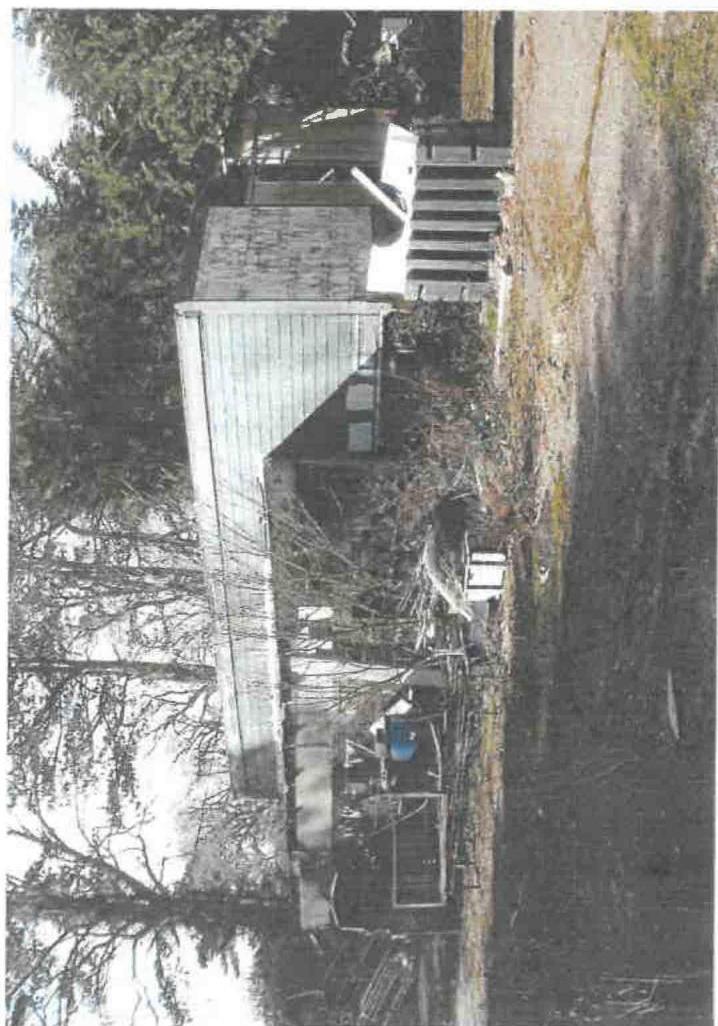 A rotated photograph showing a wooden outbuilding or shed with a pitched roof, surrounded by trees and vegetation.