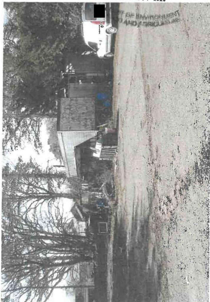 A rotated photograph showing a rural site with wooden outbuildings, a white van, and gravel ground, likely evidencing current storage use.