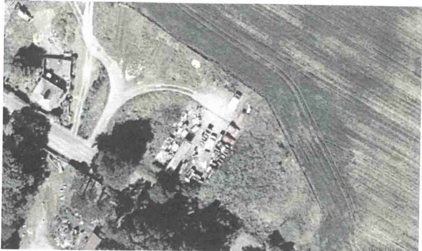 Aerial black and white photograph showing a rural site with a building complex, road junction, and adjacent agricultural field.