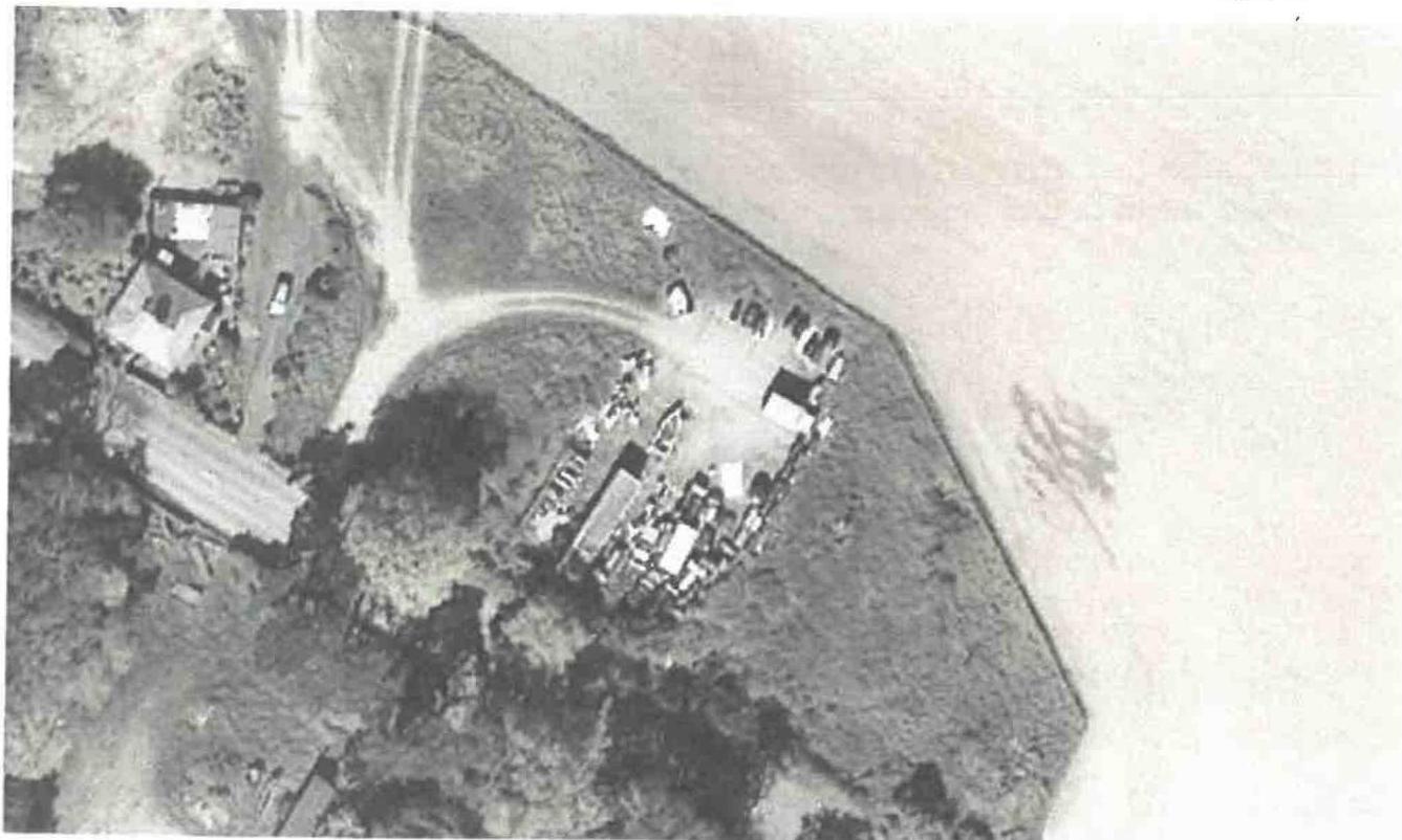 A grainy black and white aerial photograph depicting a rural plot with buildings, vehicles, and a nearby road.