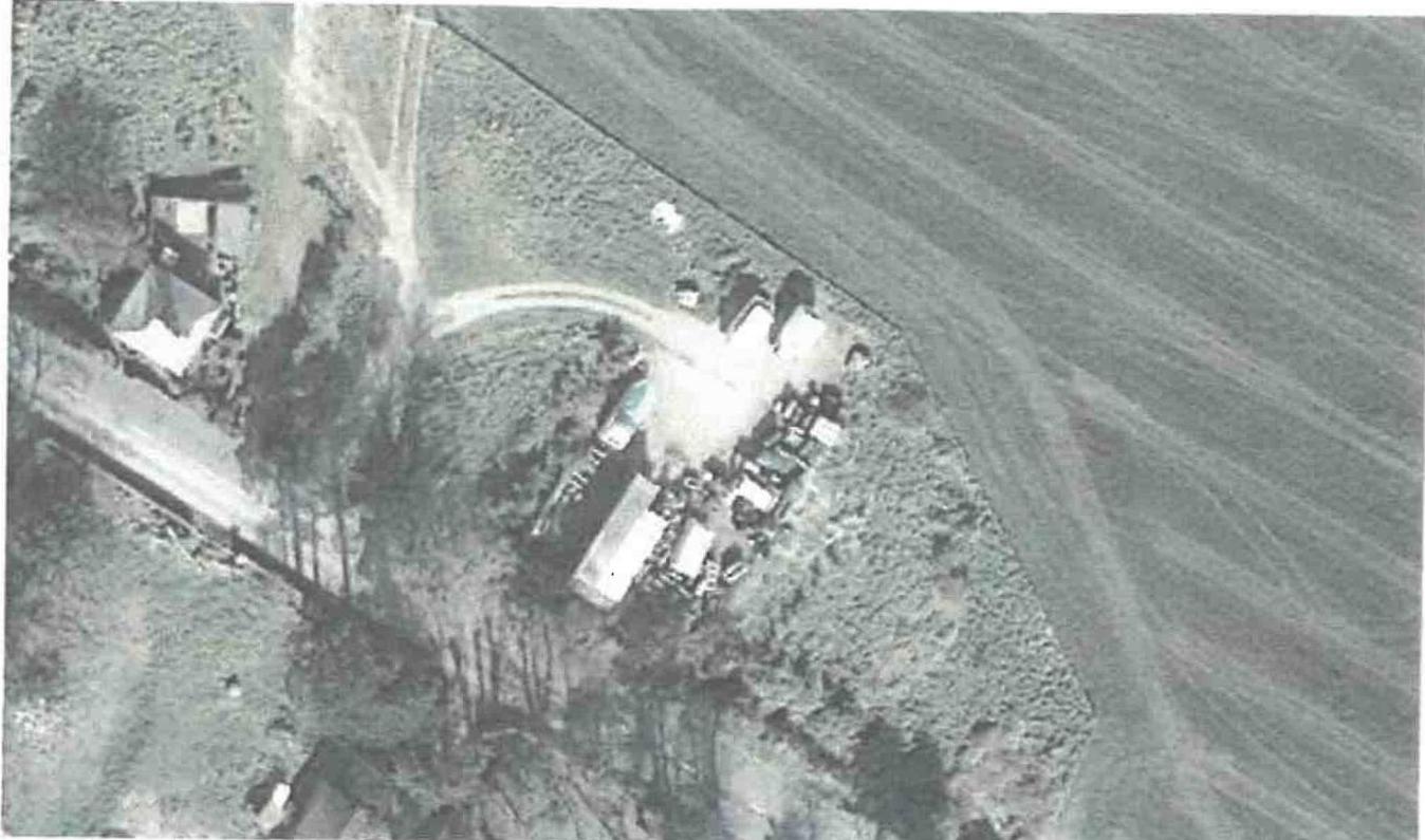 A grainy black and white aerial photograph showing a rural site with clustered buildings, tracks, and surrounding agricultural fields.