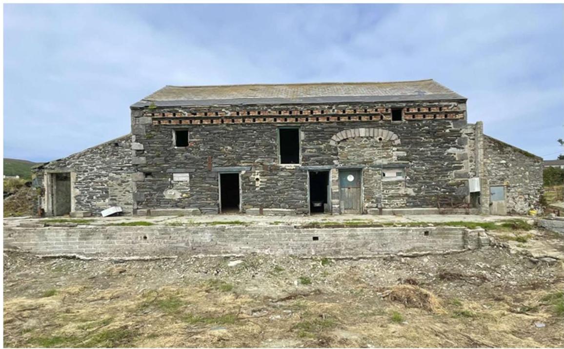 A photograph of a large, two-storey stone agricultural barn with empty window openings and exposed brickwork, appearing to be in a state of disrepair or renovation.