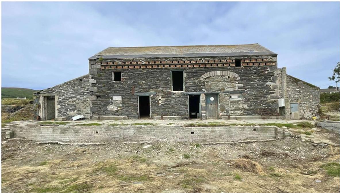 A photograph showing a large, two-story stone agricultural barn in a rural setting, with a concrete retaining wall in the foreground.
