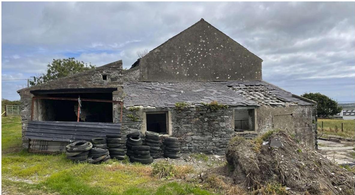 A photograph showing a dilapidated stone agricultural barn with a damaged roof and piles of tires in the foreground.
