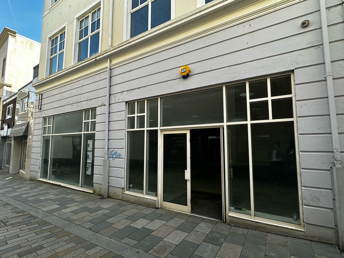 A street-level photograph showing the front elevation of a commercial building with large glass shop windows, a central entrance, and paved pavement.