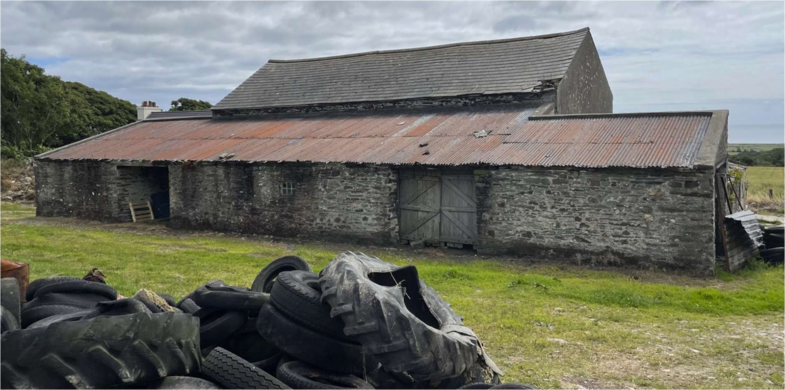 A photograph of a long, single-story stone agricultural building with a mixed slate and corrugated iron roof, set in a rural environment with debris in the foreground.
