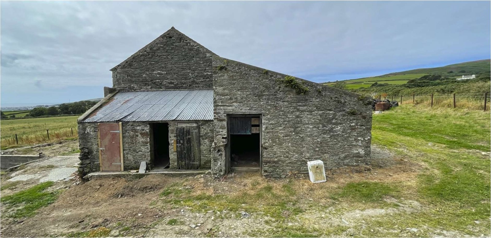 A photograph of a weathered stone agricultural barn with a corrugated metal lean-to extension situated in a rural field.