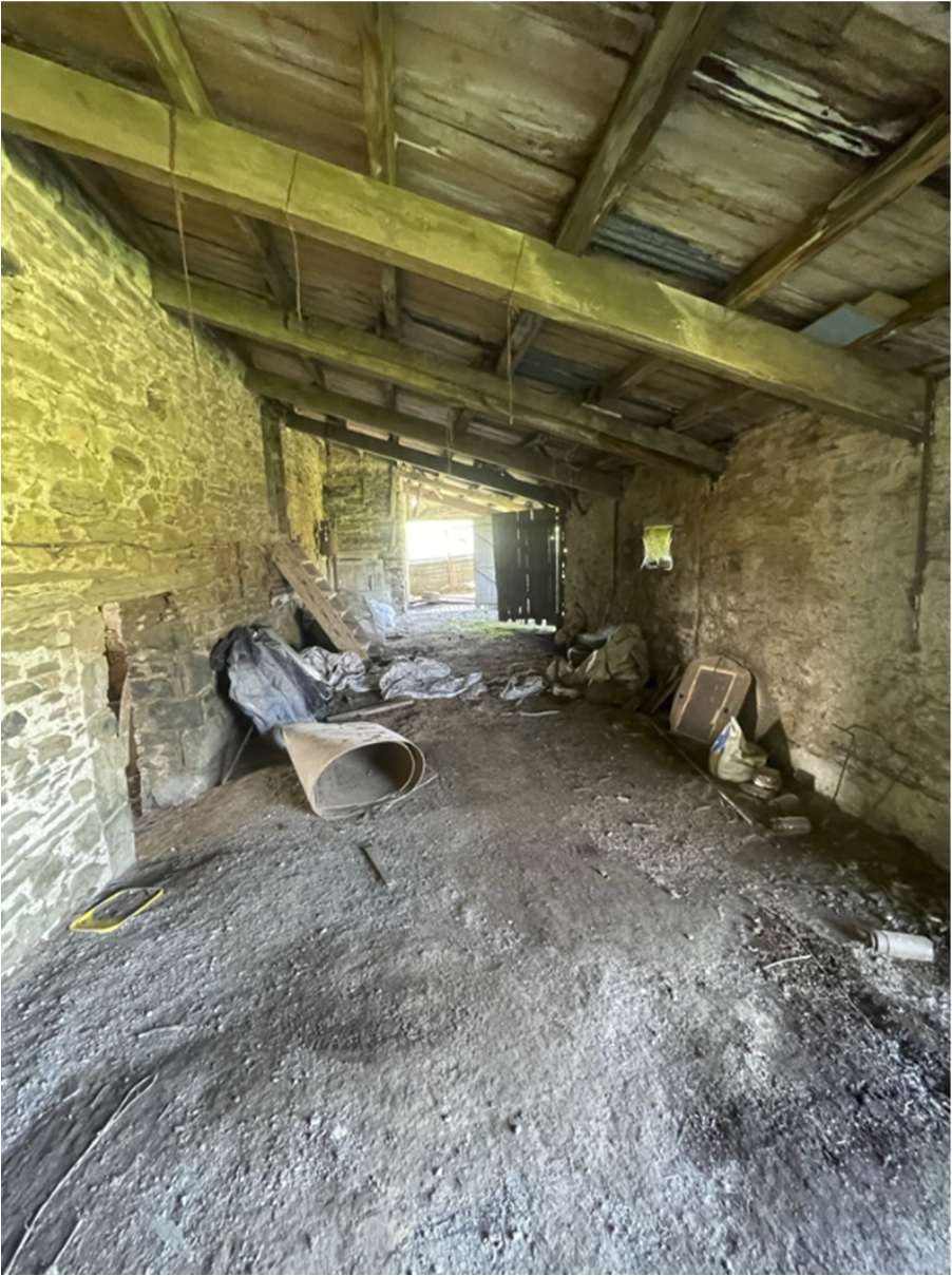 Interior photograph of a derelict agricultural barn featuring rough stone walls and exposed wooden roof beams with debris on the floor.