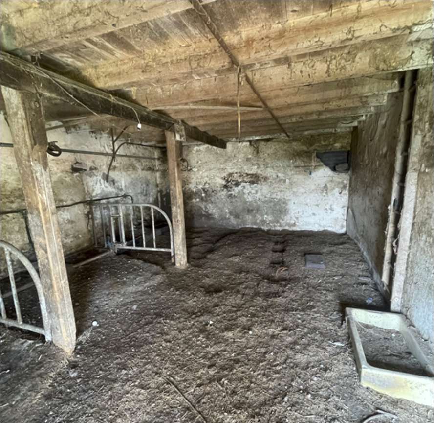 Interior view of an old agricultural barn featuring exposed wooden beams, stone walls, metal livestock gates, and a concrete feeding trough.