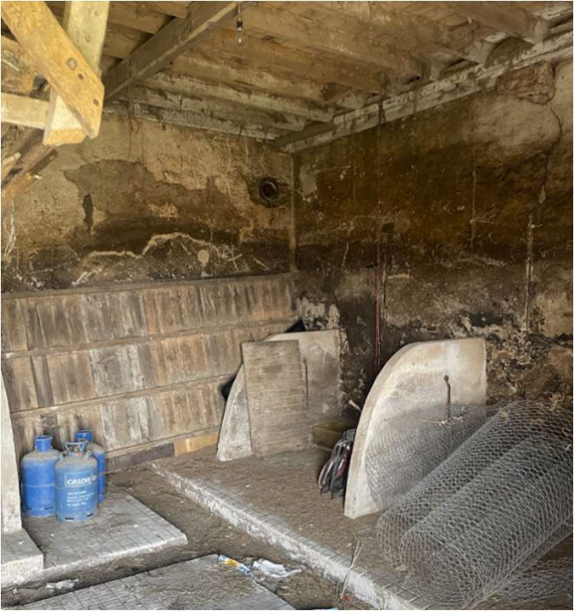 Interior photograph of a dilapidated agricultural barn showing exposed timber roof beams, rough stone walls, and stored materials like gas canisters and wire mesh.