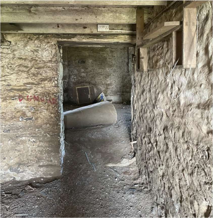 Interior photograph of a stone barn showing rough walls, wooden beams, and a doorway leading to an adjacent room.