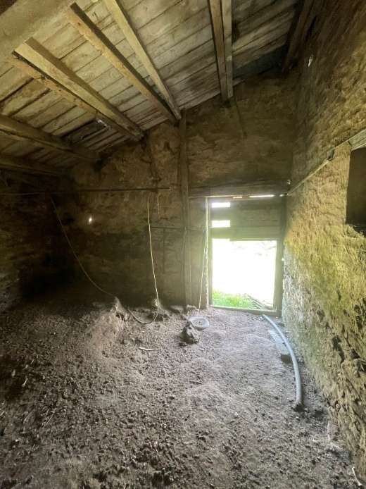 Interior photograph of an existing agricultural barn showing rough stone walls, wooden roof beams, and an earth floor.