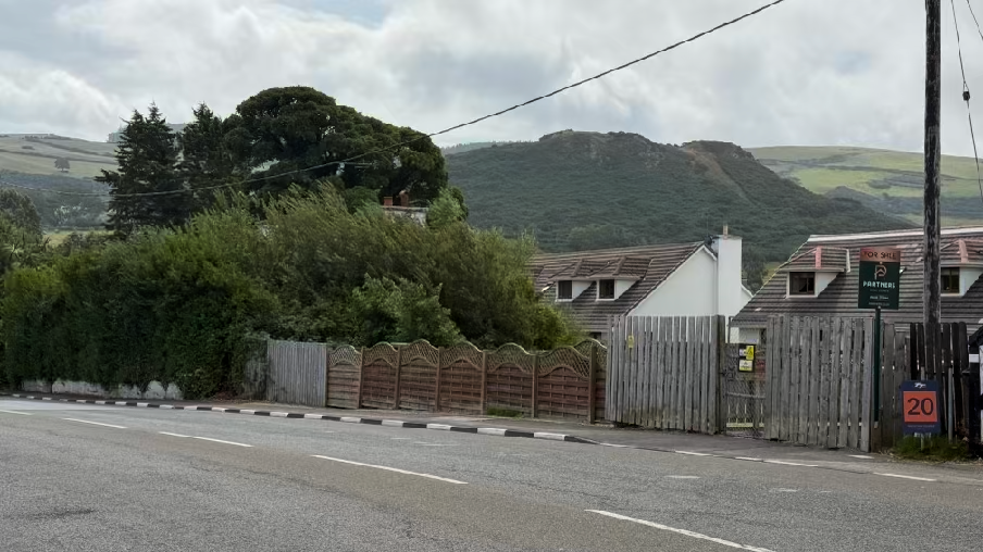 A street-level photograph showing existing residential properties behind a wooden fence, with green hills in the background.