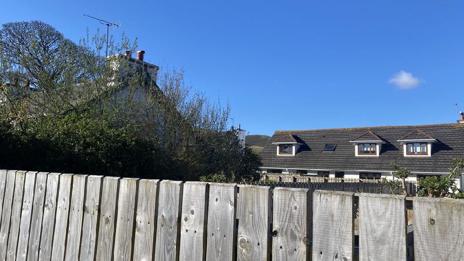 A photograph showing a wooden fence in the foreground with residential houses visible in the background, including a house with dormer windows.