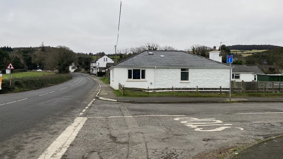 A white single-story bungalow situated at a road junction in a rural setting, featuring a grey slate roof and wooden fencing.